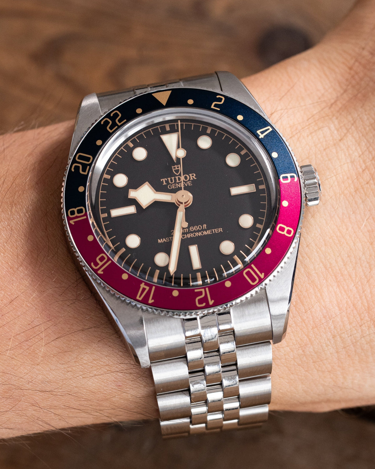 A Tudor watch with a black dial, silver stainless steel bracelet, and a two-tone blue and red bezel, shown worn on a wrist against a wooden background.