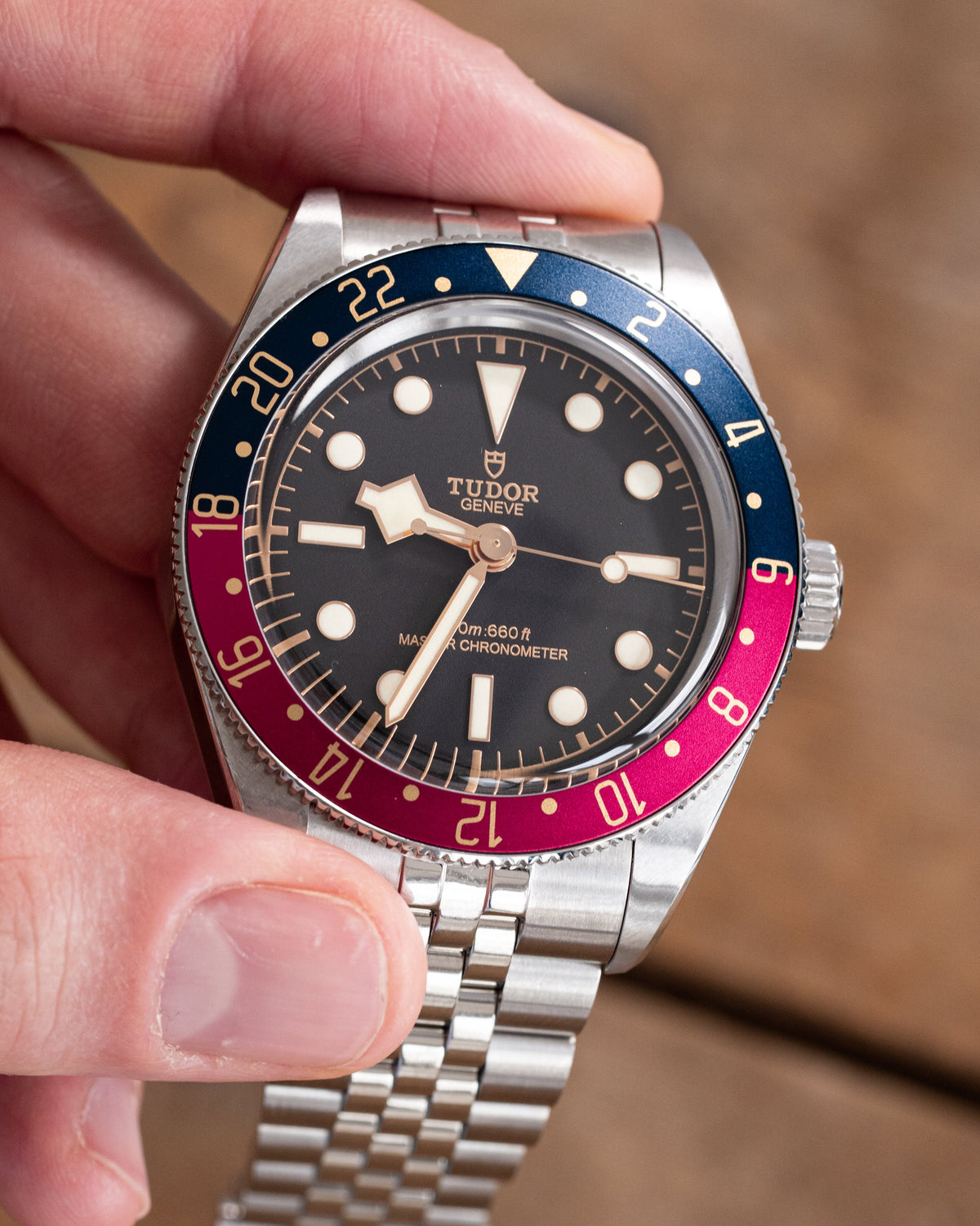A close-up of a hand holding a Tudor watch with a silver stainless steel bracelet and a two-tone blue and red bezel against a warm brown background.