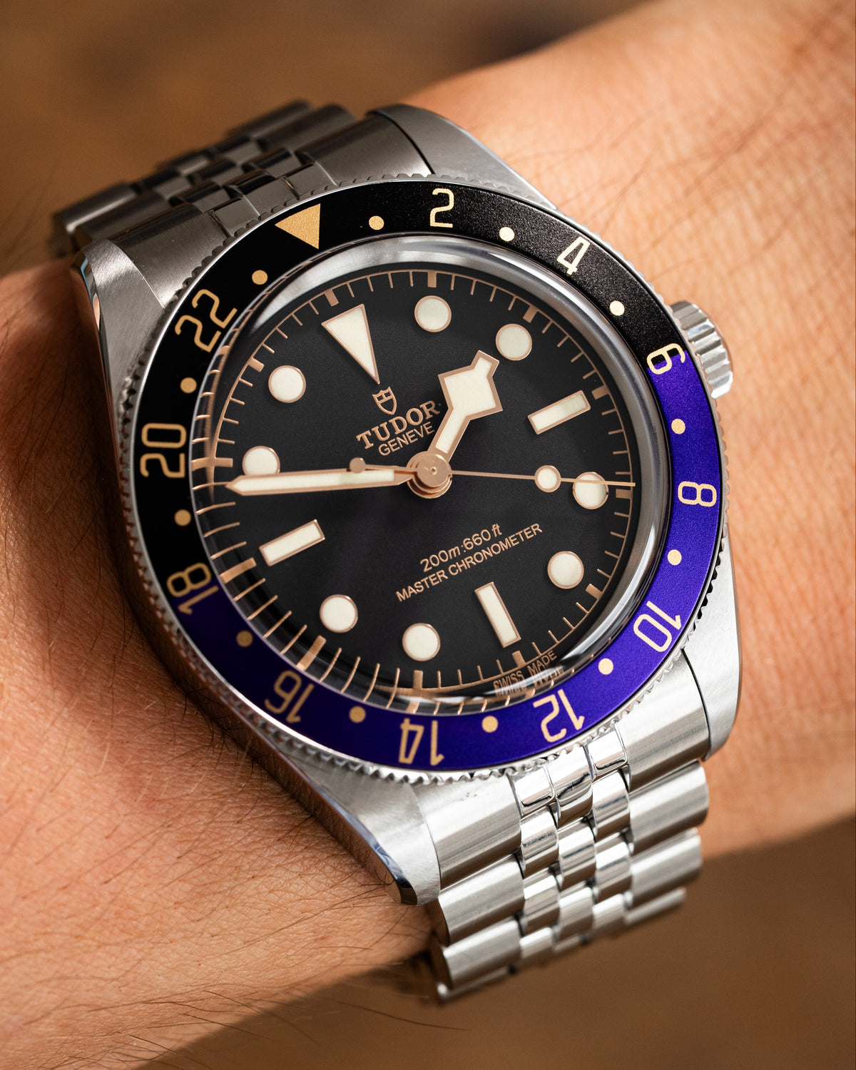 A close-up of a wrist wearing a Tudor watch with a silver stainless steel bracelet, black dial, and a purple and black bezel.
