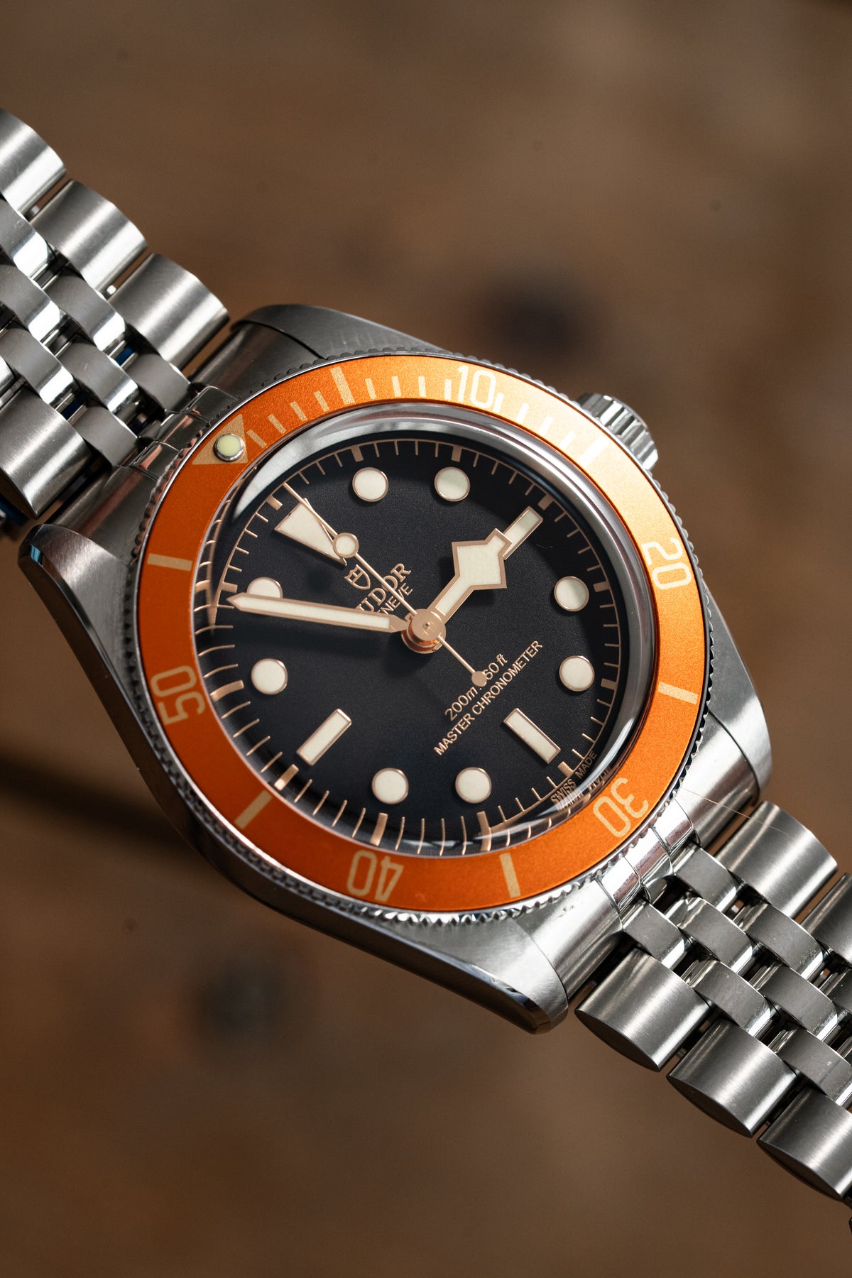 A close-up shot of a Tudor diver's watch with a silver stainless steel bracelet, a black dial, and a vibrant orange rotating bezel, set against a blurred brown background.