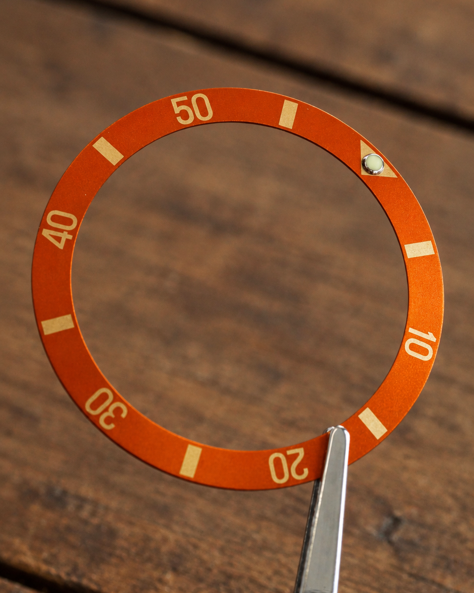 An orange circular bezel ring with white numbers 10, 20, 30, 40, 50 and white indicator marks, held by metal tweezers on a dark wooden surface.