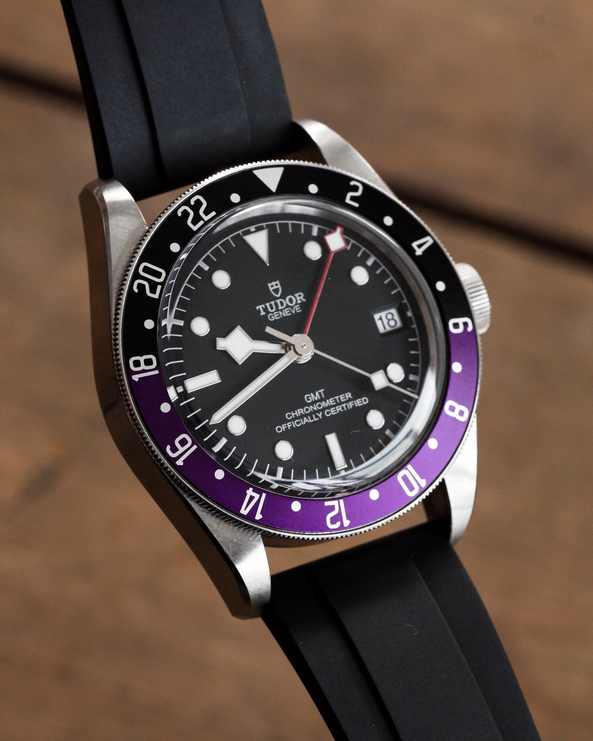 A close-up of a Tudor GMT watch with a black rubber strap, black dial, and a bezel featuring a purple lower section and white upper section, resting on a wooden surface.