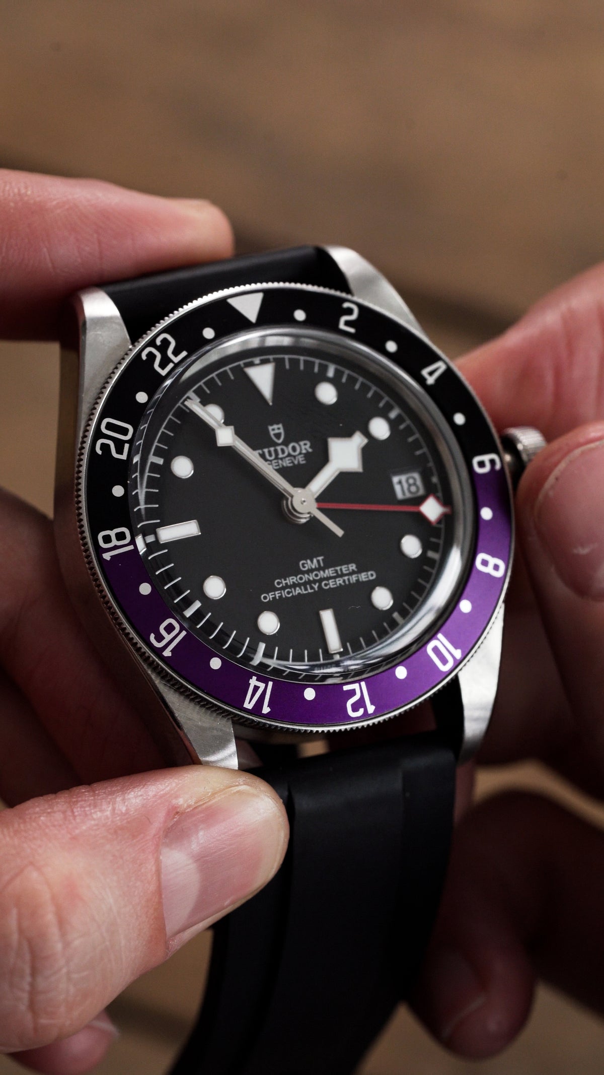 A close-up of a Tudor Black Bay GMT wristwatch held in hands, featuring a black dial, a black and purple bezel, and a black rubber strap.