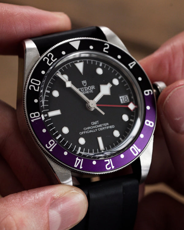 A close-up of a Tudor Black Bay GMT wristwatch held in hands, featuring a black dial, a black and purple bezel, and a black rubber strap.